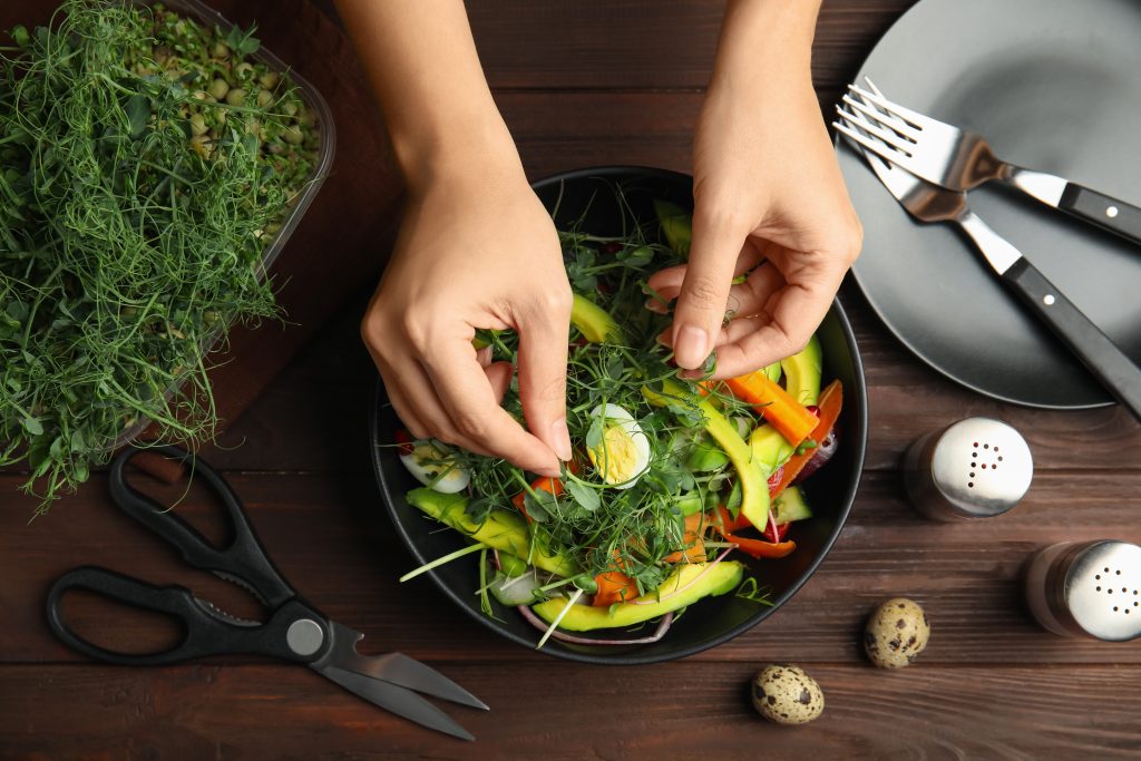 Woman making salad with fresh organic microgreens at wooden table, top view
